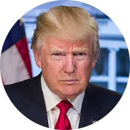 Portrait of a man with light hair wearing a black suit and red tie, with a blurred American flag and building background.